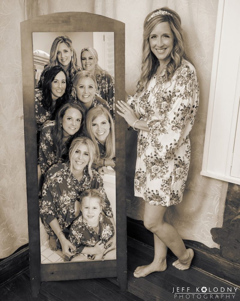 Bride standing next to mirror showing reflection of bridesmaids - Picture by Jeff Kolodny Photography