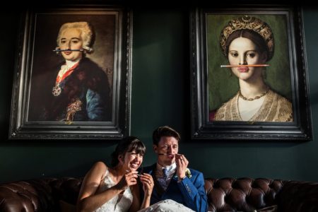 Bride laughing as groom puts pen under his nose like faces in pictures behind them - Picture by Rafe Abrook Photography