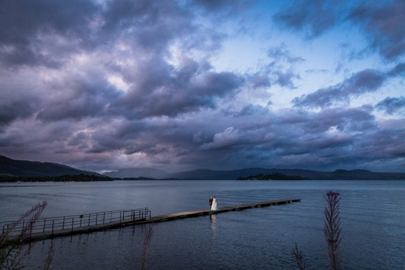 Bride and groom walking across long jetty under cloudy sky - Picture by First Light Photography