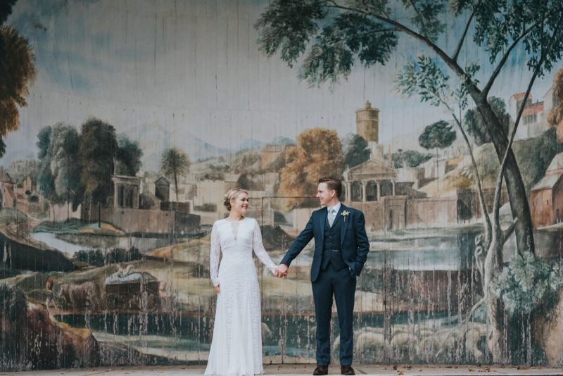 Bride and groom holding hands in front of rustic backdrop - Picture by Matt Fox Photography