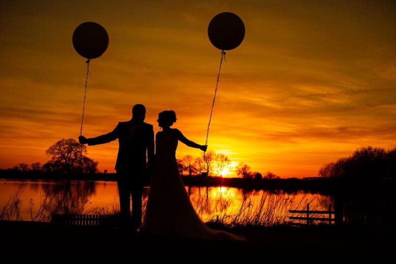 Silhouette of bride and groom holding balloons against sunset - Picture by Carpe Diem Photography