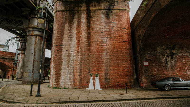Two brides against wall holding bouquets up their faces - Picture by Maddie Farris Photography