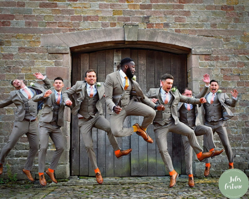 Groomsmen in grey suits and brown shoes jumping in the air - Picture by Jules Fortune Photography