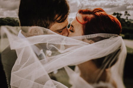 Bride and groom kissing behind veil blowing in wind - Picture by Oobaloos Photography