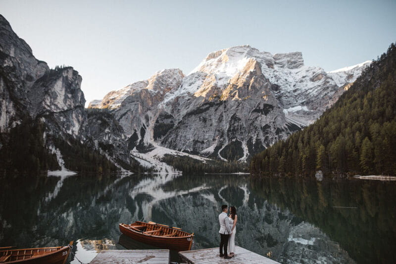 Bride and groom standing by lake with snow-covered mountains in background - Picture by Katja & Simon Photography