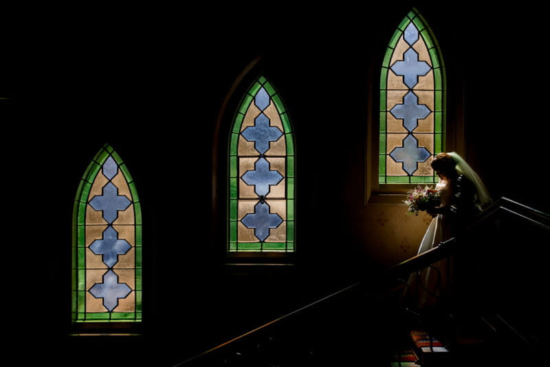 Bride walking down stairs past stained glass windows - Picture by Damion Mower Photography