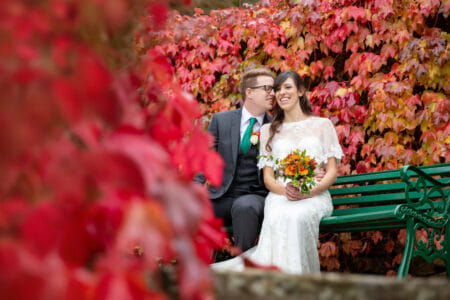 Bride and groom sitting on bench by bushes with red leaves - Picture by Helen England Photography
