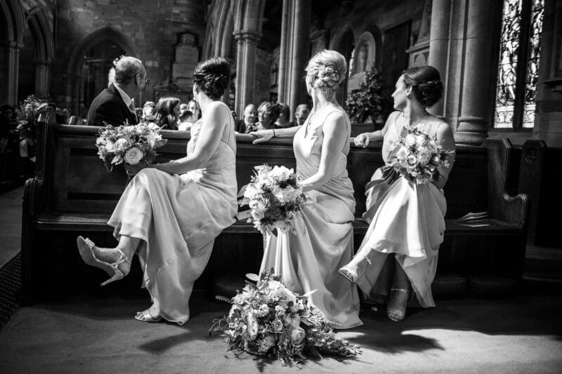 Three bridesmaids sitting in church looking over their shoulders at the same time - Picture by Nick Brightman Photography