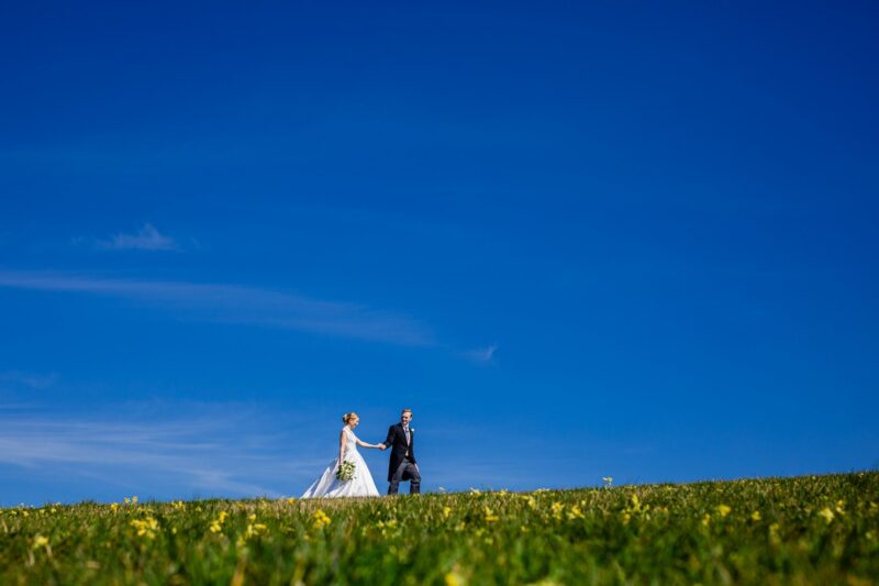 Bride and groom walking on grass underneath bright blue sky - Picture by How Photography