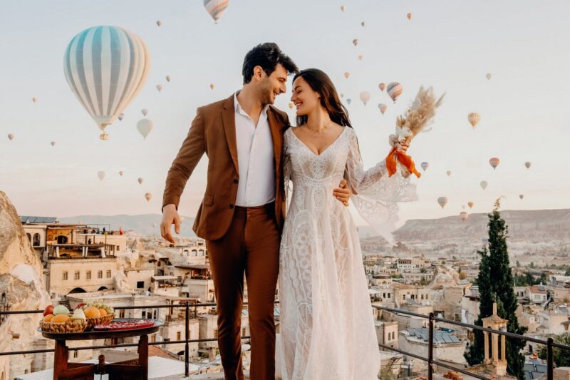 Bride and groom standing on balcony with hot air balloons in sky behind them - Picture by Emma-Jane Photography