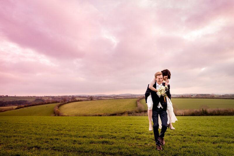 Groom giving bride piggy back ride across field with pink sky overhead - Picture by Lee Maxwell Photography