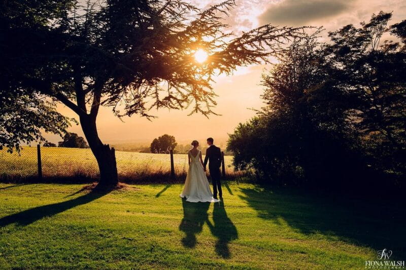 Bride and groom holding hands by tree as sun sets - Picture by Fiona Walsh Photography