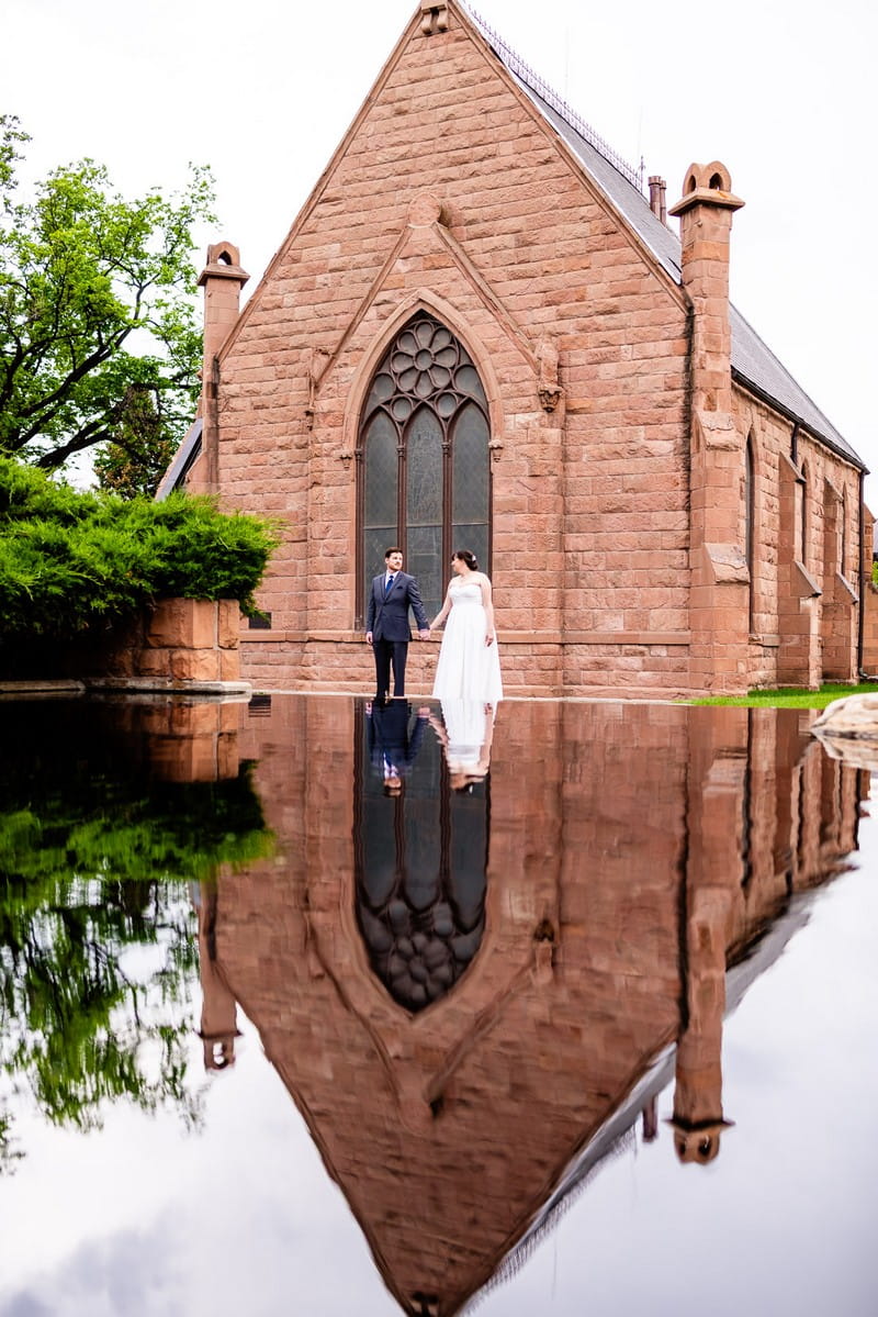 Bride and groom holding hands outside church with their reflection showing in water - Picture by Gabrielle Stowe Photography