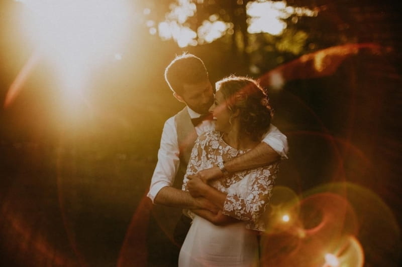 Groom hugging bride from behind as sun shines through trees - Picture by Jason Mark Harris