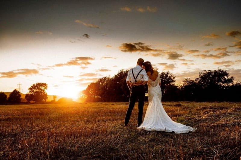 Bride and groom standing in field watching sun set - Picture by Weddings by Tom Langford